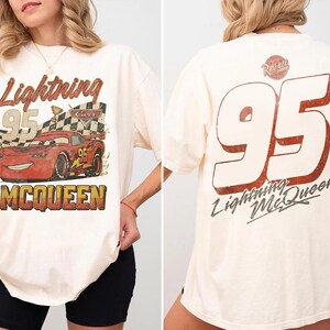 Op de afbeelding: Cr&egrave;mekleurig T-shirt met een versleten afbeelding van Lightning McQueen uit de film Cars. De voorkant toont het personage met de tekst "Lightning 95 McQueen". De achterkant heeft een grote "95" en de tekst "Lightning McQueen".