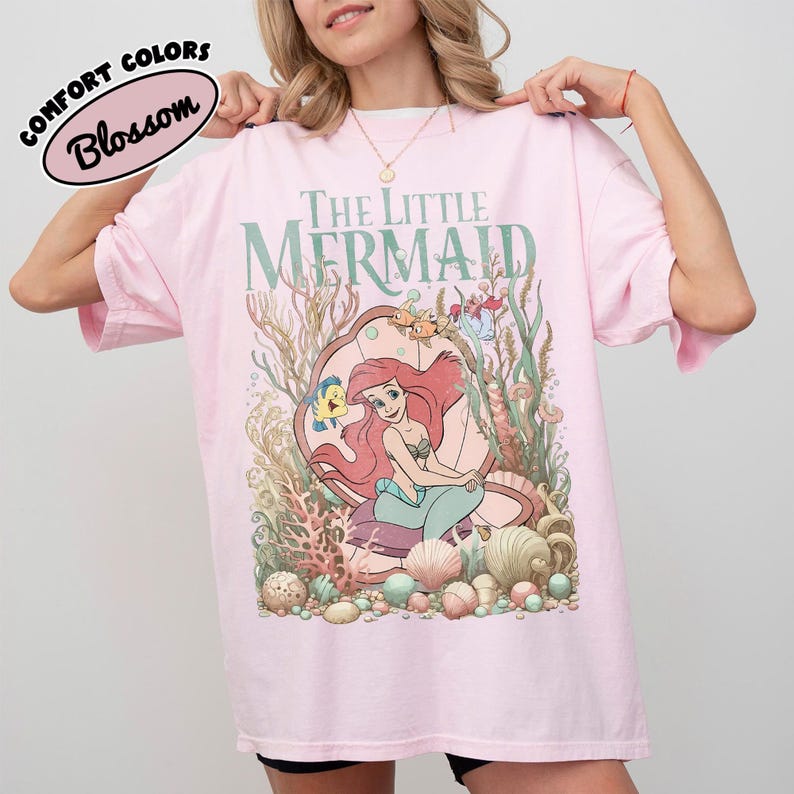 May include: A light pink t-shirt featuring the text "THE LITTLE MERMAID" in green. The shirt has a graphic of Ariel, Flounder, and Sebastian. The words "COMFORT COLORS" and "Blossom" are in a white oval on the top left.