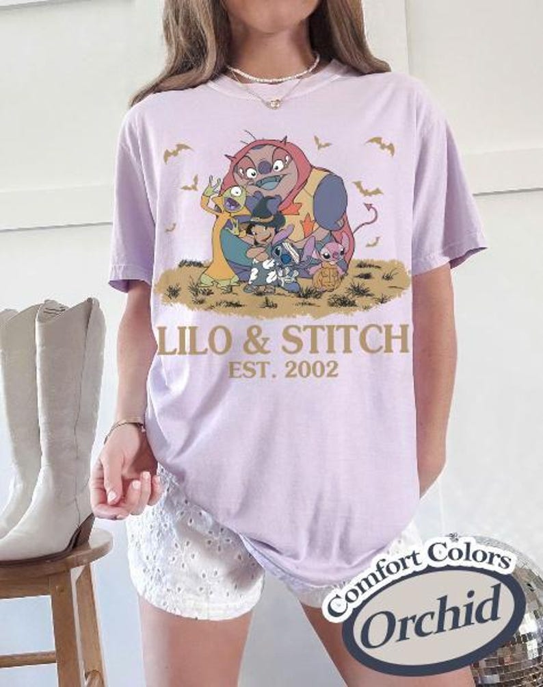 Op de afbeelding: Lichtpaars T-shirt met een afbeelding van Lilo & Stitch-personages en de tekst "Lilo & Stitch EST. 2002". Het shirt is een Comfort Colors Orchid. Het ontwerp bevat stripfiguren en vleermuizen.