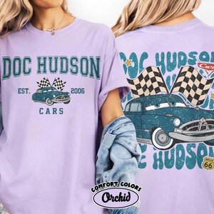 May include: A light purple t-shirt featuring a graphic of Doc Hudson's car from the movie Cars. The front of the shirt has the text "DOC HUDSON CARS EST. 2006" above the car image. The back of the shirt has a similar design.