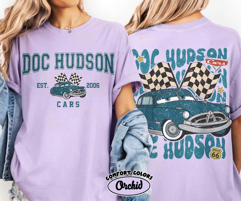 May include: A light purple t-shirt featuring a graphic of Doc Hudson's car from the movie Cars. The front of the shirt has the text "DOC HUDSON CARS EST. 2006" above the car image. The back of the shirt has a similar design.