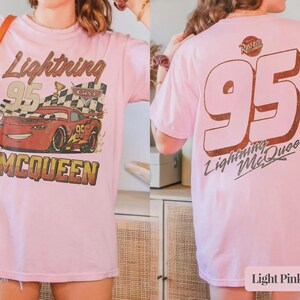 Op de afbeelding: Lichtroze T-shirt met een Lightning McQueen-afbeelding uit de film Cars. De voorkant toont de tekst "Lightning 95 McQueen" met een cartoonauto en een geblokte vlag. De achterkant van het shirt heeft een grote "95" en de tekst "Lightning McQueen".
