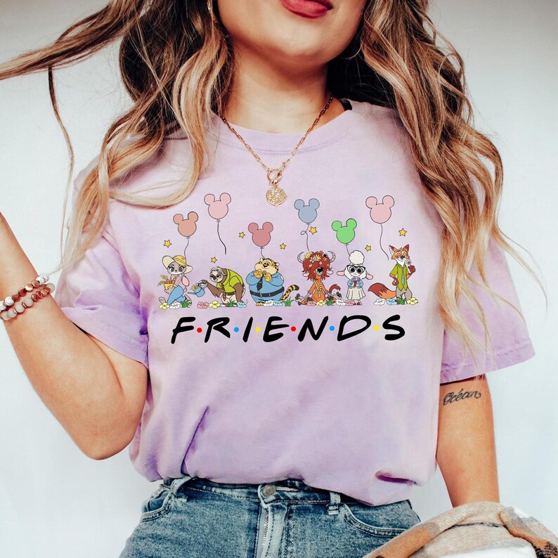 May include: A light purple t-shirt featuring cartoon characters holding Mickey Mouse-shaped balloons. The word "FRIENDS" is printed in black block letters below the characters. The shirt is a crew neck style.