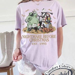 Puede incluir: Una camiseta lila con un gr&aacute;fico de "Pesadilla antes de Navidad". El dise&ntilde;o incluye personajes como Jack Skellington, Sally y Oogie Boogie, con el texto "NIGHTMARE BEFORE CHRISTMAS EST. 1993". La camiseta es un Comfort Colors Orchid.