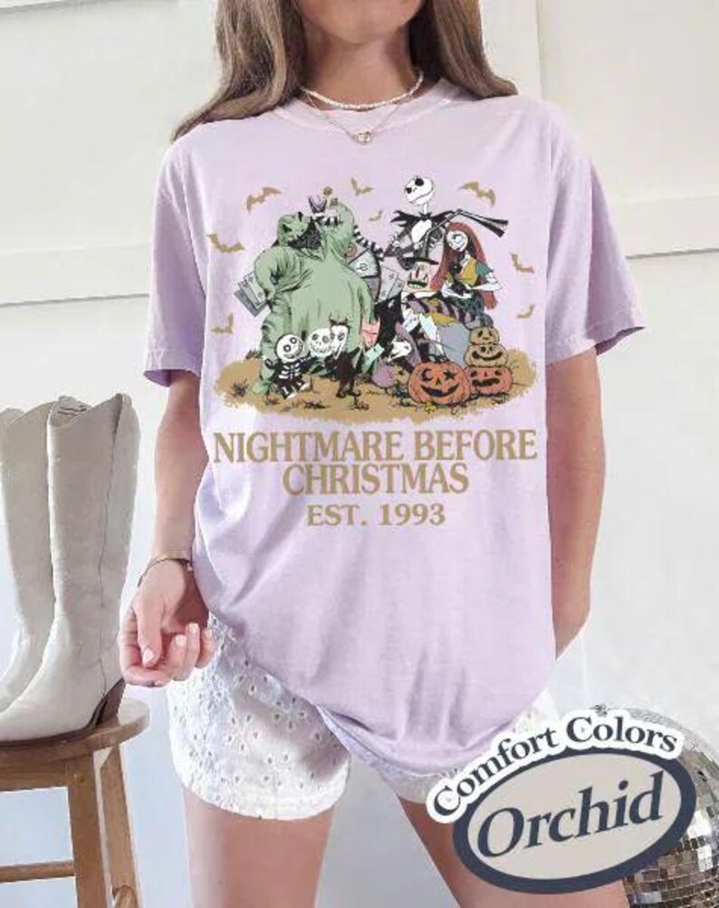 Puede incluir: Una camiseta lila con un gr&aacute;fico de "Pesadilla antes de Navidad". El dise&ntilde;o incluye personajes como Jack Skellington, Sally y Oogie Boogie, con el texto "NIGHTMARE BEFORE CHRISTMAS EST. 1993". La camiseta es un Comfort Colors Orchid.