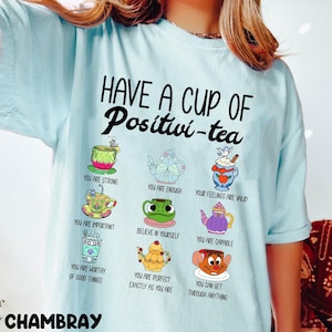 Puede incluir: Camiseta azul claro con el texto "HAVE A CUP OF Positivi-tea" y varias tazas de té de dibujos animados con afirmaciones positivas. La camiseta presenta la palabra "CHAMBRAY" en la parte inferior izquierda.