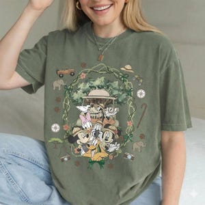 Puede incluir: Camiseta verde oliva con un dise&ntilde;o tem&aacute;tico de safari. El dise&ntilde;o incluye a Mickey Mouse, Minnie Mouse, Donald Duck, Goofy y Daisy Duck con atuendos de safari, rodeados de elementos de safari como un jeep, una br&uacute;jula y binoculares.