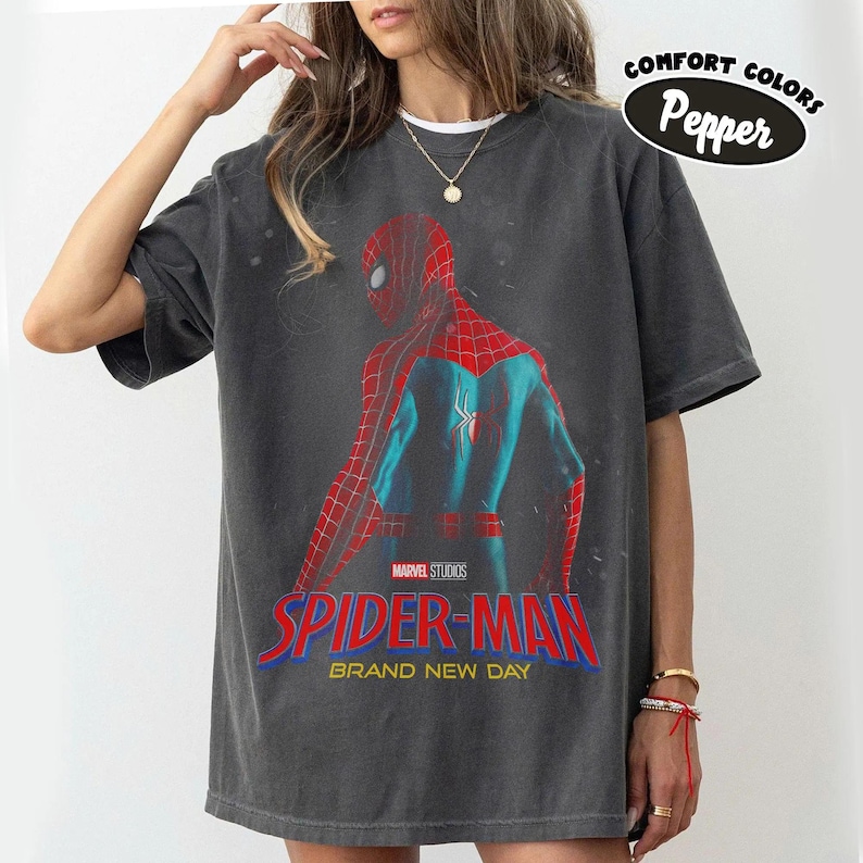 May include: Dark grey t-shirt with a Spider-Man graphic from Marvel Studios. The design shows Spider-Man's back in red and blue. The text "SPIDER-MAN BRAND NEW DAY" is displayed in red and blue. The shirt also has the text "Comfort Colors Pepper".