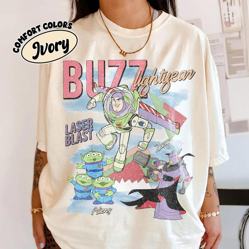 Puede incluir: Camiseta de color marfil con un gr&aacute;fico de Buzz Lightyear, extraterrestres y Zurg de la pel&iacute;cula Toy Story. El dise&ntilde;o incluye las palabras "BUZZ lightyear" y "LASER BLAST" en rosa y azul. La camiseta es de la marca Comfort Colors.