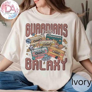 May include: Ivory-colored t-shirt featuring the text "GUARDIANS of the GALAXY" in a retro font. The design includes cassette tapes with phrases like "Hooked on a feeling" and "Spirit in the sky," along with planets and stars.
