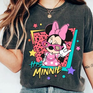 K&ouml;nnte beinhalten: Dunkelgraues T-Shirt mit einer Minnie Mouse-Grafik, die eine rosa Sonnenbrille und ein gepunktetes Kleid tr&auml;gt. Das Design enth&auml;lt den Text "His Minnie" in Gelb, vor einem farbenfrohen geometrischen Hintergrund mit Sternen.