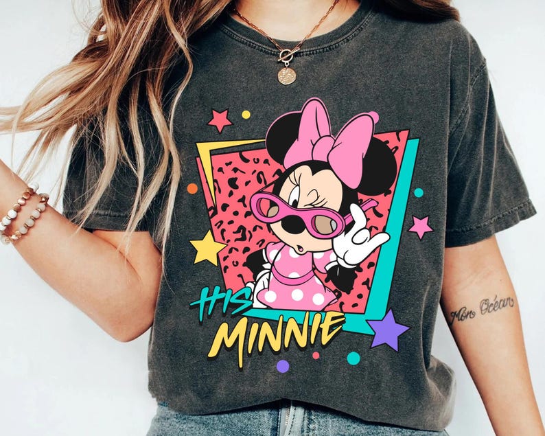 K&ouml;nnte beinhalten: Dunkelgraues T-Shirt mit einer Minnie Mouse-Grafik, die eine rosa Sonnenbrille und ein gepunktetes Kleid tr&auml;gt. Das Design enth&auml;lt den Text "His Minnie" in Gelb, vor einem farbenfrohen geometrischen Hintergrund mit Sternen.
