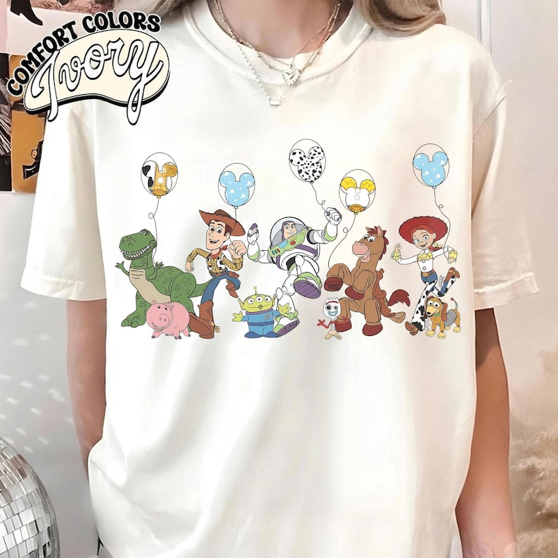 K&ouml;nnte beinhalten: Cremefarbenes T-Shirt mit einer bunten Grafik von Zeichentrickfiguren, die Luftballons halten. Die Figuren umfassen einen gr&uuml;nen Dinosaurier, ein Schwein, einen Cowboy, einen Astronauten, einen Au&szlig;erirdischen, ein Pferd und ein Cowgirl. Das Shirt hat den Schriftzug "Comfort Colors Ivory".