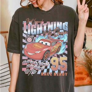 May include: Dark gray t-shirt featuring a graphic of Lightning McQueen from the movie Cars. The design includes the text "Street Legend Lightning McQueen", "Need for Speed", and "Race Ready". The number 95 is prominently displayed.