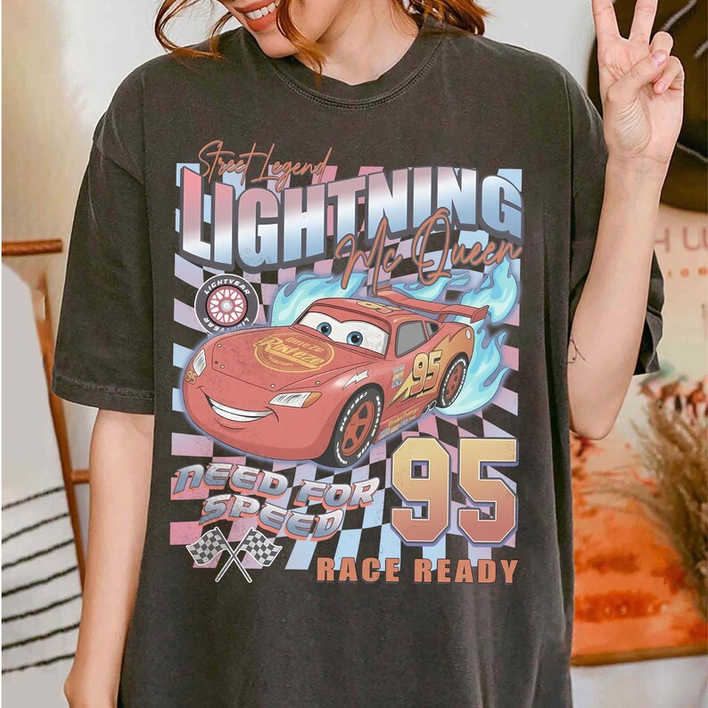 May include: Dark gray t-shirt featuring a graphic of Lightning McQueen from the movie Cars. The design includes the text "Street Legend Lightning McQueen", "Need for Speed", and "Race Ready". The number 95 is prominently displayed.
