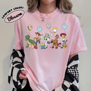 K&ouml;nnte beinhalten: Hellrosa Comfort Colors T-Shirt mit Toy Story-Charakteren, die Ballons halten. Zu den Charakteren geh&ouml;ren Rex, Woody, Buzz Lightyear und Jessie. Das Shirt hat das Wort "Blossom" in einem kreisf&ouml;rmigen Design.