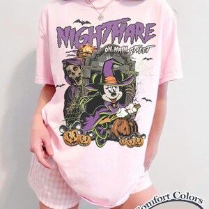 Op de afbeelding: Zachtroze T-shirt met een Halloween-thema. Het ontwerp toont Mickey Mouse in een heksenkostuum, pompoenen en de tekst "Nightmare on Main Street". Het shirt is gelabeld "Comfort Colors Blossom".