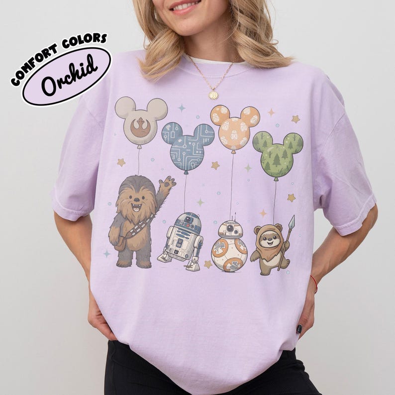 May include: Orchid Comfort Colors t-shirt featuring Star Wars characters holding Mickey Mouse-shaped balloons. Chewbacca, R2-D2, BB-8, and an Ewok are depicted. The balloons have various patterns. The shirt is a light purple color.