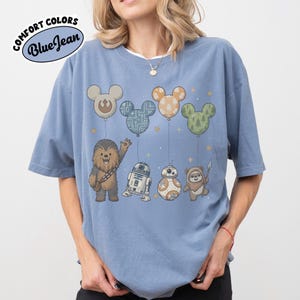 May include: Blue t-shirt with a graphic featuring Star Wars characters and Mickey Mouse-shaped balloons. The shirt has the text "Comfort Colors Blue Jean" in a circular design. The characters include Chewbacca, R2-D2, BB-8, and an Ewok.