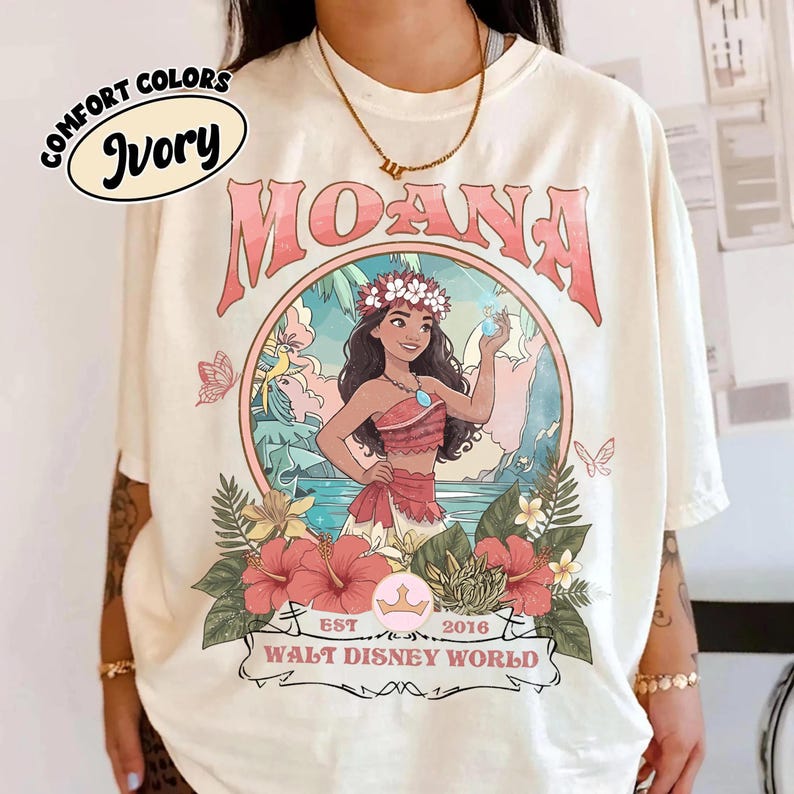 May include: Ivory-colored t-shirt featuring a circular graphic of Moana, surrounded by flowers and tropical scenery. The word "MOANA" is arched above the image. The shirt also includes the text "EST 2016 WALT DISNEY WORLD" in a banner.