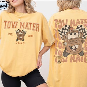 May include: Mustard-colored t-shirt featuring the text "TOW MATER" above a cartoon image of Tow Mater from the movie Cars, with checkered flags. The back of the shirt repeats the design. The shirt is a comfort color.