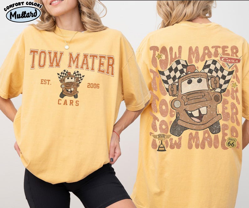 May include: Mustard-colored t-shirt featuring the text "TOW MATER" above a cartoon image of Tow Mater from the movie Cars, with checkered flags. The back of the shirt repeats the design. The shirt is a comfort color.