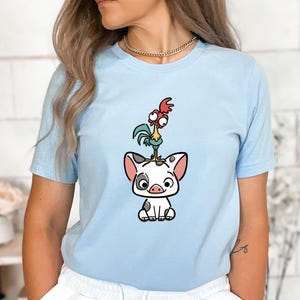 Puede incluir: Camiseta azul claro con una ilustraci&oacute;n de dibujos animados de un cerdo con manchas negras, un gallo y una gallina. El cerdo est&aacute; sentado y el gallo est&aacute; de pie sobre la cabeza del cerdo. La gallina est&aacute; de pie sobre la cabeza del gallo.