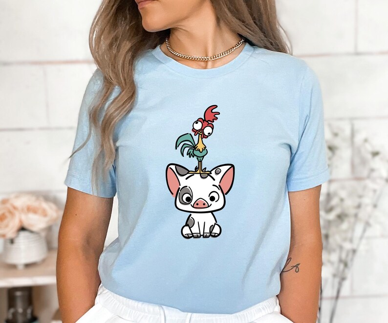 Puede incluir: Camiseta azul claro con una ilustraci&oacute;n de dibujos animados de un cerdo con manchas negras, un gallo y una gallina. El cerdo est&aacute; sentado y el gallo est&aacute; de pie sobre la cabeza del cerdo. La gallina est&aacute; de pie sobre la cabeza del gallo.