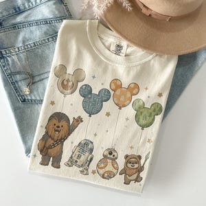 May include: Cream-colored t-shirt featuring Star Wars characters and Mickey Mouse-shaped balloons. Chewbacca, R2-D2, BB-8, and an Ewok are depicted. The balloons have various patterns and colors, with small star accents.