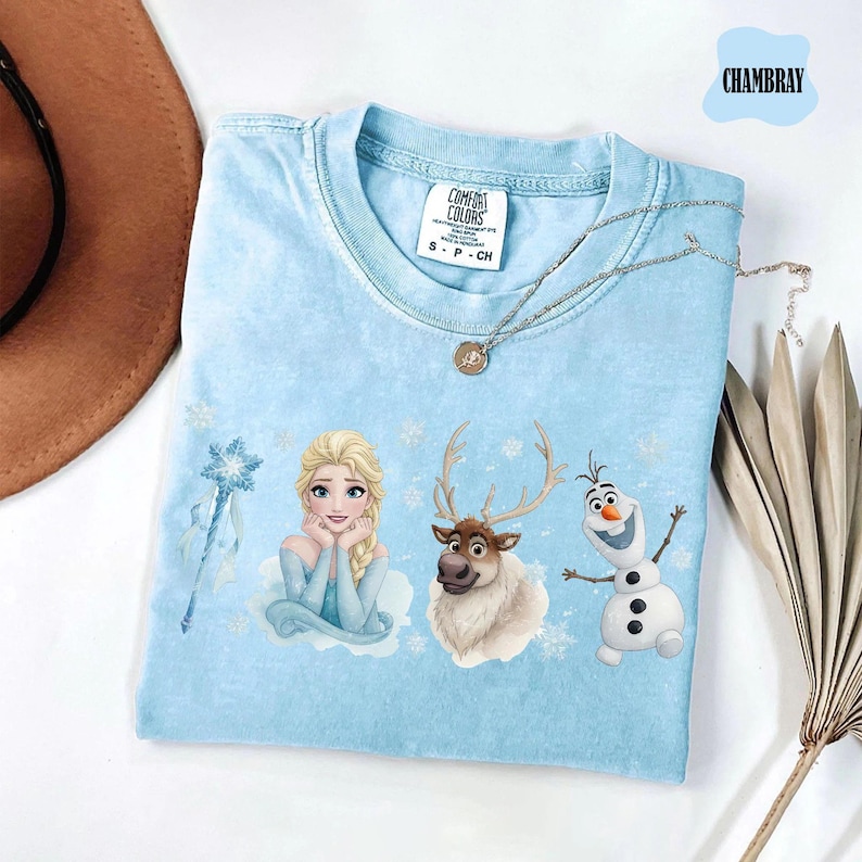 Peut inclure: T-shirt bleu clair avec Elsa, un renne et Olaf de La Reine des Neiges. Le t-shirt a un col rond et des manches courtes. Un collier argent&eacute; avec un pendentif est drap&eacute; sur le t-shirt. Le t-shirt est &eacute;tiquet&eacute; "Chambray".