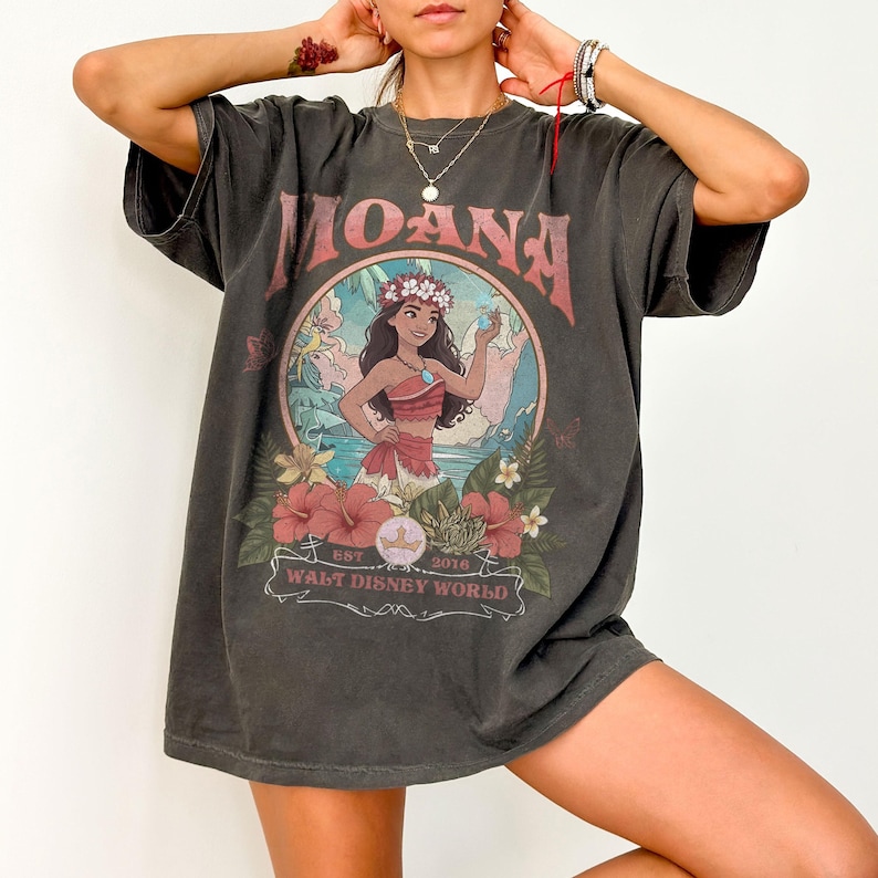 May include: Dark gray t-shirt featuring the word "MOANA" in a pink, stylized font. The shirt has a circular graphic of a cartoon character surrounded by flowers and the text "WALT DISNEY WORLD".