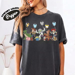 K&ouml;nnte beinhalten: Dunkelgraues T-Shirt mit einer farbenfrohen Grafik von Toy Story-Charakteren, die Ballons halten. Der Text "COMFORT COLORS Pepper" ist oben links aufgedruckt. Das Design umfasst Woody, Buzz Lightyear und andere Charaktere.