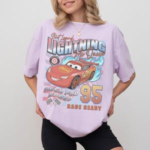 May include: A light purple t-shirt featuring a graphic of Lightning McQueen from the movie Cars. The design includes the text "Street Legend Lightning Mc Queen", "Need for Speed", and "Race Ready", along with the number "95".