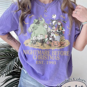 Puede incluir: Camiseta violeta con un dise&ntilde;o gr&aacute;fico de "Pesadilla antes de Navidad". El dise&ntilde;o incluye personajes como Jack Skellington y Sally, con el texto "NIGHTMARE BEFORE CHRISTMAS EST. 1993."