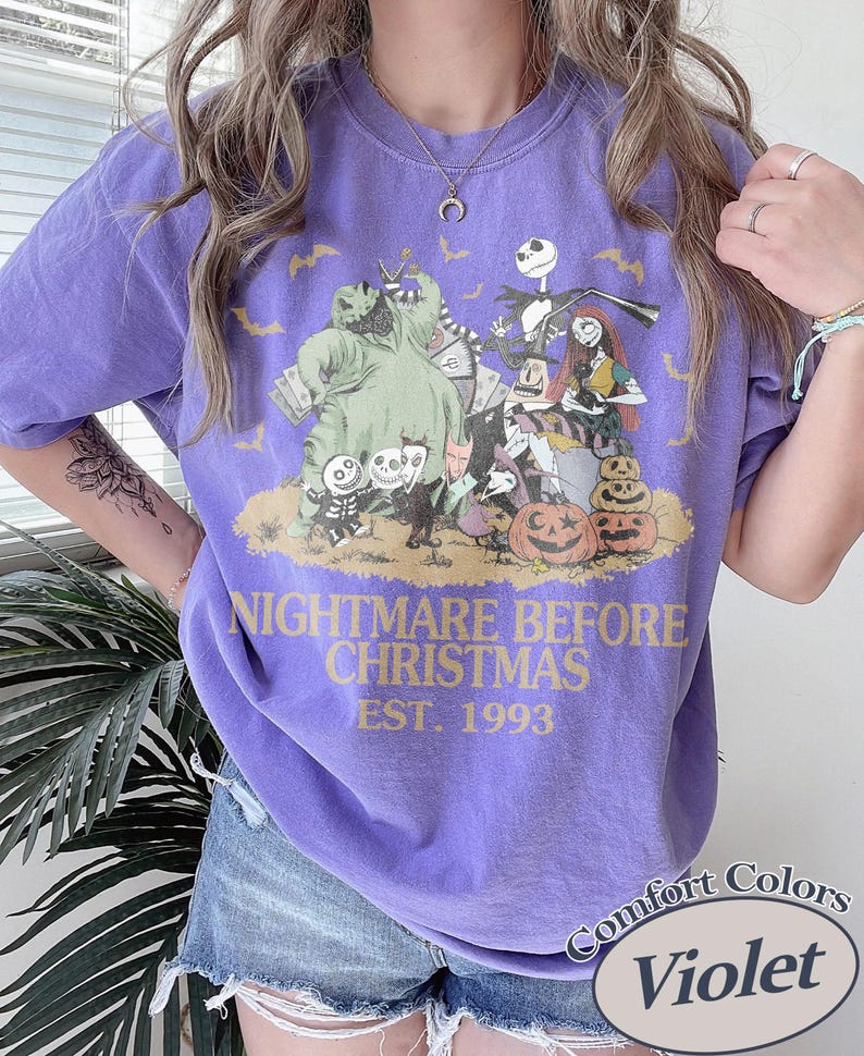 Puede incluir: Camiseta violeta con un dise&ntilde;o gr&aacute;fico de "Pesadilla antes de Navidad". El dise&ntilde;o incluye personajes como Jack Skellington y Sally, con el texto "NIGHTMARE BEFORE CHRISTMAS EST. 1993."