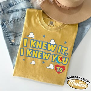 Puede incluir: Una camiseta color amarillo mostaza con el texto azul I KNEW IT, I KNEW YOU, un coraz&oacute;n rojo con las letras TS y un gr&aacute;fico de pelota de colores. La camiseta tiene cuello redondo y una etiqueta de la marca Comfort Colors en el cuello.
