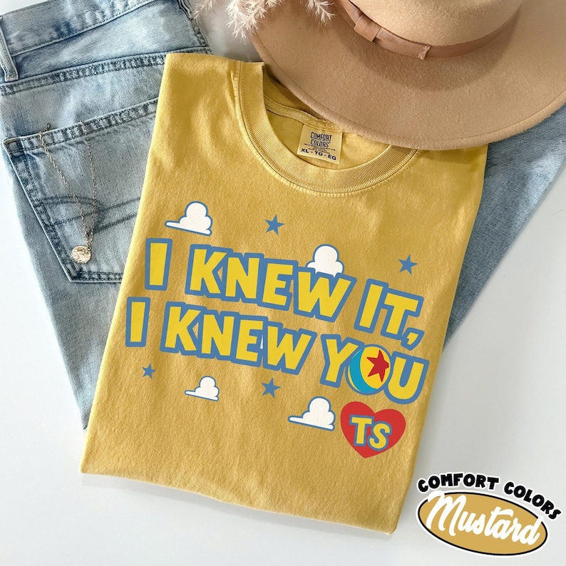 Puede incluir: Una camiseta color amarillo mostaza con el texto azul I KNEW IT, I KNEW YOU, un coraz&oacute;n rojo con las letras TS y un gr&aacute;fico de pelota de colores. La camiseta tiene cuello redondo y una etiqueta de la marca Comfort Colors en el cuello.