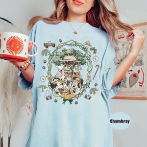 Puede incluir: Camiseta azul claro con un dise&ntilde;o tem&aacute;tico de safari con Mickey Mouse, el Pato Donald y otros personajes de Disney. El dise&ntilde;o incluye un jeep, una br&uacute;jula y huellas de patas. La palabra "Chambray" est&aacute; impresa en la parte inferior derecha.
