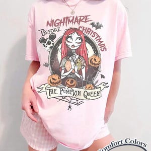May include: A light pink t-shirt featuring a graphic of Sally from The Nightmare Before Christmas surrounded by jack-o'-lanterns, bats, and a skull. The text reads Nightmare Before Christmas and The Pumpkin Queen. A Comfort Colors Blossom label is visible.