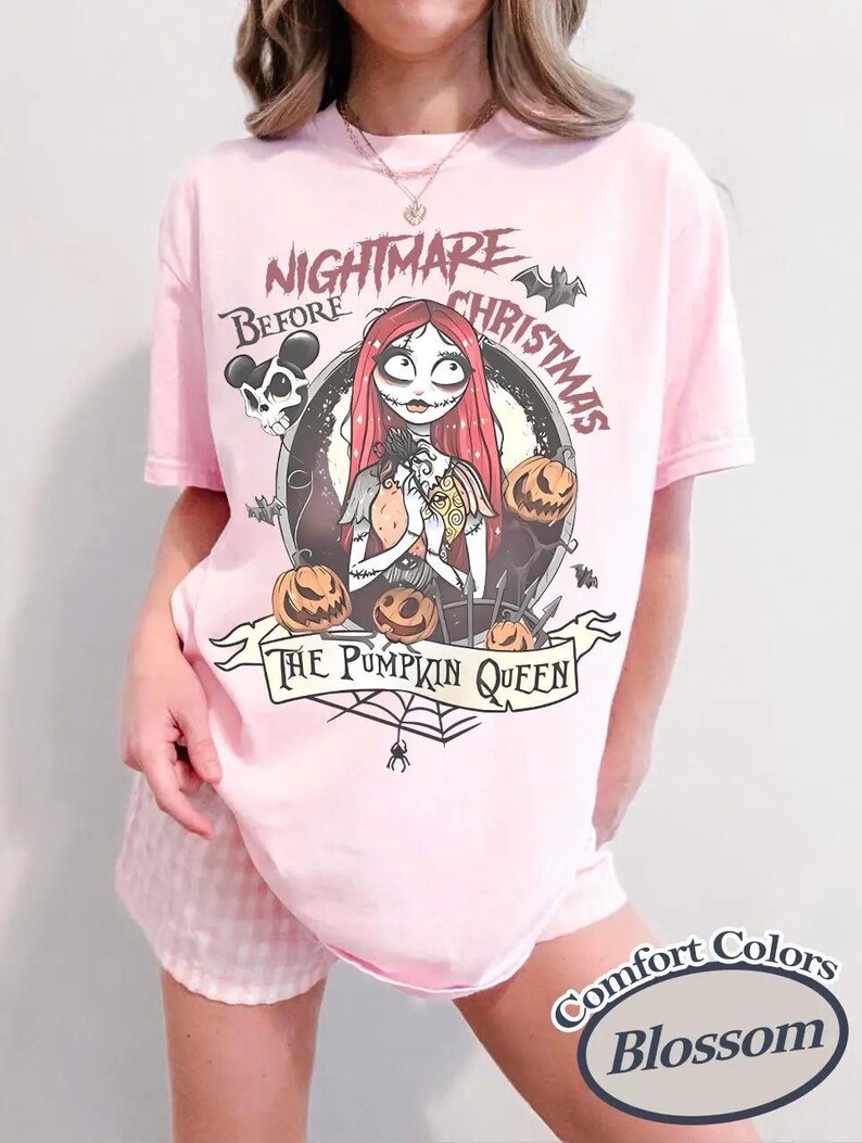 May include: A light pink t-shirt featuring a graphic of Sally from The Nightmare Before Christmas surrounded by jack-o'-lanterns, bats, and a skull. The text reads Nightmare Before Christmas and The Pumpkin Queen. A Comfort Colors Blossom label is visible.