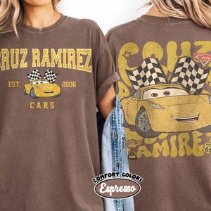 May include: Brown t-shirt featuring a graphic of Cruz Ramirez from the Cars movie. The front design includes the text "CRUZ RAMIREZ" above a yellow car with checkered flags, and "EST. 2006 CARS" below. The back design repeats the car graphic and text.