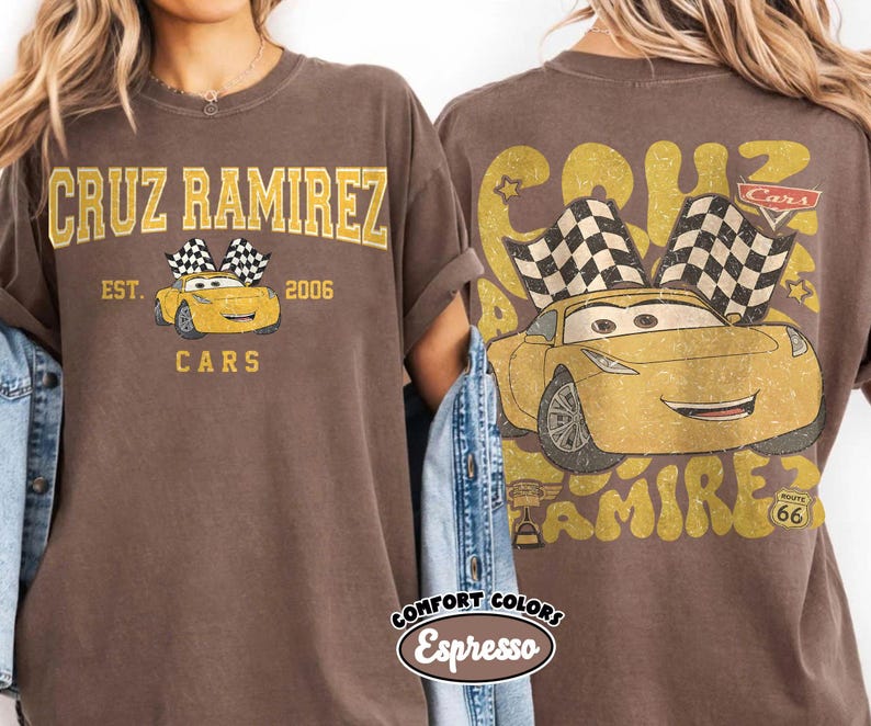 May include: Brown t-shirt featuring a graphic of Cruz Ramirez from the Cars movie. The front design includes the text "CRUZ RAMIREZ" above a yellow car with checkered flags, and "EST. 2006 CARS" below. The back design repeats the car graphic and text.