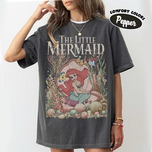 May include: Dark gray t-shirt featuring the text "The Little Mermaid" above an illustration of Ariel in an underwater scene with Flounder and other sea creatures. The shirt has a Comfort Colors Pepper label.