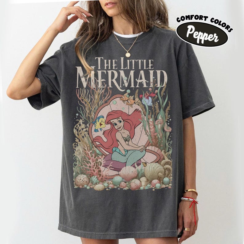 May include: Dark gray t-shirt featuring the text "The Little Mermaid" above an illustration of Ariel in an underwater scene with Flounder and other sea creatures. The shirt has a Comfort Colors Pepper label.