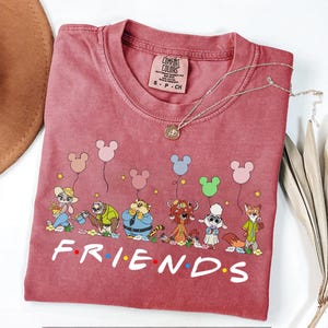 May include: A maroon Comfort Colors t-shirt featuring a cartoon design of Disney characters holding Mickey Mouse balloons. The word "FRIENDS" is printed in white at the bottom. A silver necklace with a pendant is draped on the shirt.
