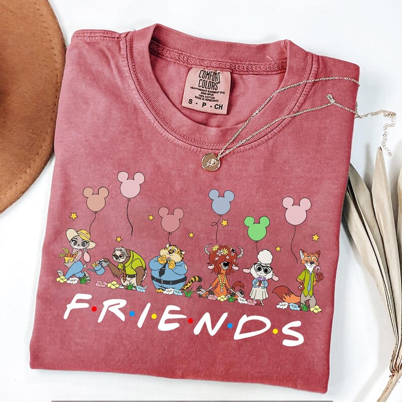 May include: A maroon Comfort Colors t-shirt featuring a cartoon design of Disney characters holding Mickey Mouse balloons. The word "FRIENDS" is printed in white at the bottom. A silver necklace with a pendant is draped on the shirt.