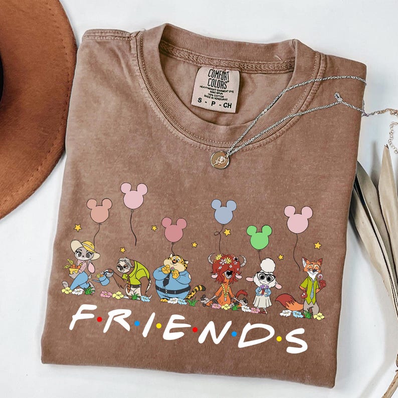 May include: Brown Comfort Colors t-shirt featuring a cartoon graphic of animal characters holding Mickey Mouse-shaped balloons. The word "FRIENDS" is printed in white block letters across the bottom. A silver necklace with a pendant is draped across the shirt.