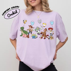 K&ouml;nnte beinhalten: Orchideenfarbenes T-Shirt mit einer Grafik von Toy Story-Charakteren, die Luftballons halten. Die Ballons haben verschiedene Designs, darunter Mickey Mouse-Ohren. Der Text "COMFORT COLORS" und "Orchid" befindet sich in einem wei&szlig;en Oval.