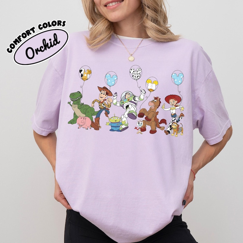 K&ouml;nnte beinhalten: Orchideenfarbenes T-Shirt mit einer Grafik von Toy Story-Charakteren, die Luftballons halten. Die Ballons haben verschiedene Designs, darunter Mickey Mouse-Ohren. Der Text "COMFORT COLORS" und "Orchid" befindet sich in einem wei&szlig;en Oval.