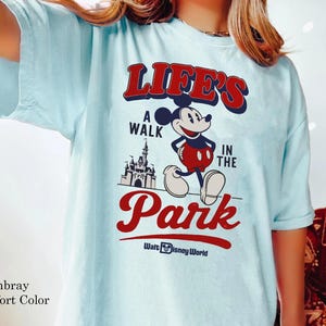 Puede incluir: Camiseta azul claro con un gr&aacute;fico de Mickey Mouse y el texto "LIFE'S A WALK IN THE PARK" en rojo y blanco. El dise&ntilde;o incluye un castillo y "Walt Disney World". La camiseta es de la marca Comfort Color.
